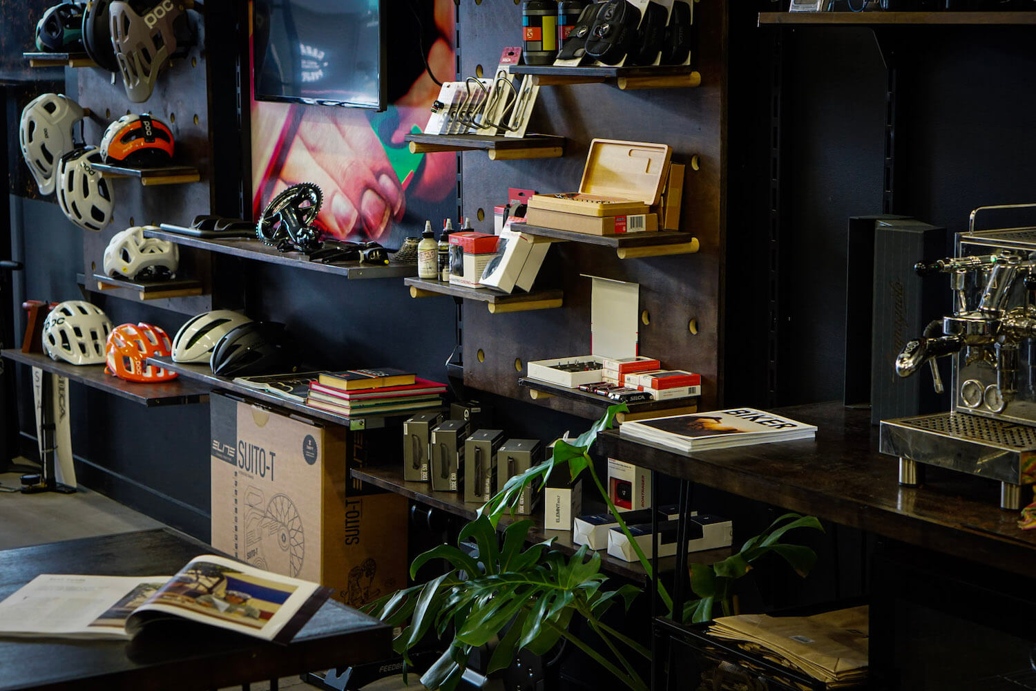 Lots of different helmets and acessories stacked on wooden benches attached to a wall with a plant in the front a box in the middle